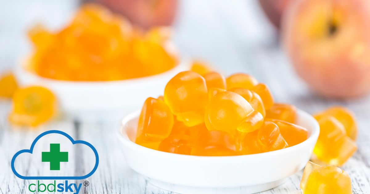 Two bowls of CBD SKY peach gummies accompanied by fresh peaches on a white wooden table.